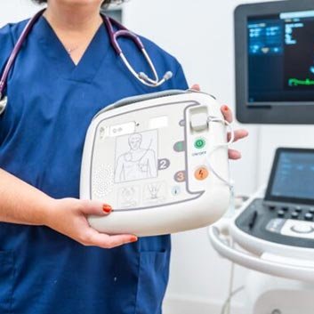 A healthcare professional in scrubs holds a defibrillator, with a medical monitor visible in the background, ready for use.