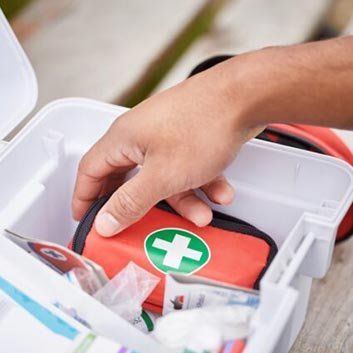 A hand reaches for a small red first aid kit inside an open white first aid box, surrounded by various medical supplies.